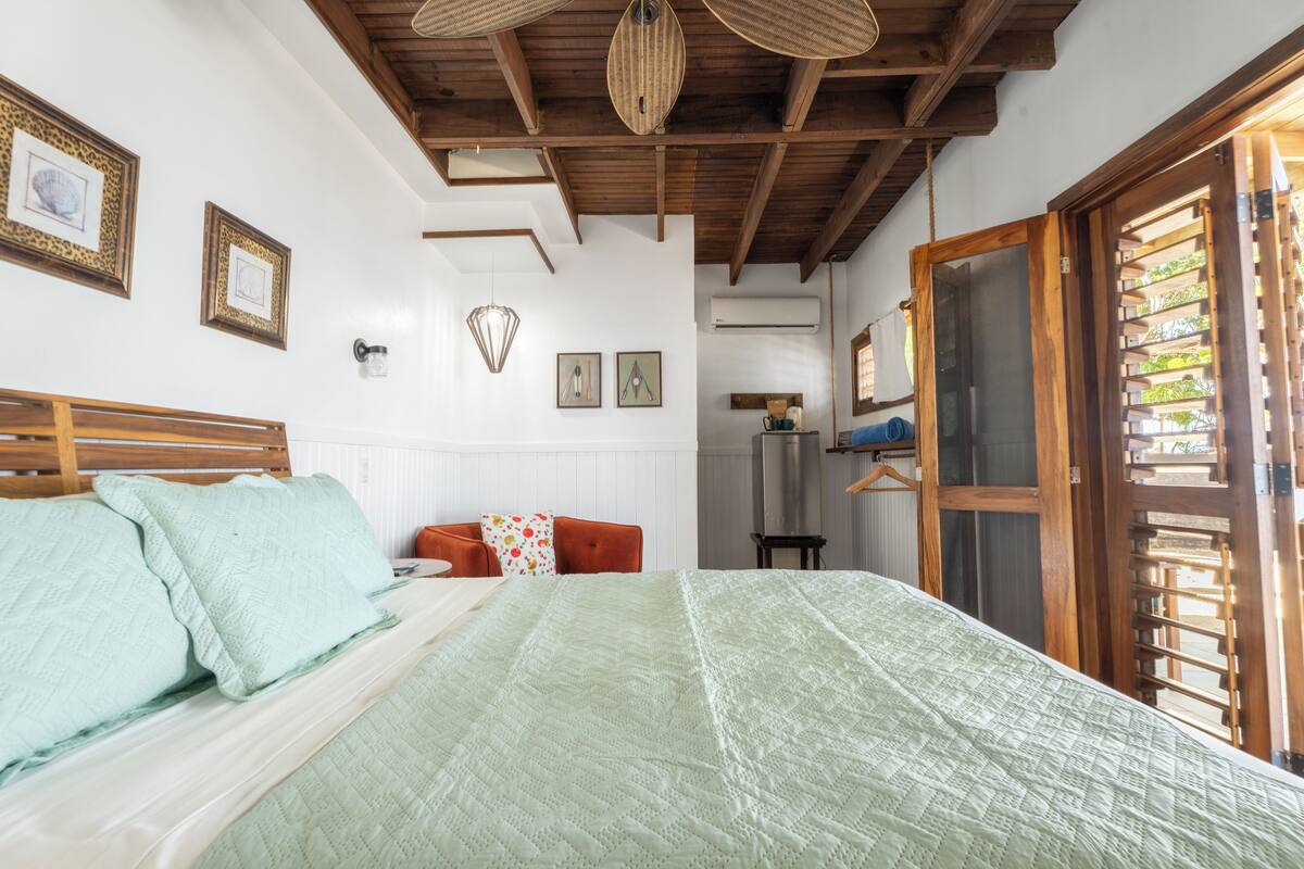 A bedroom with a large bed, wooden ceiling beams, and a view of a garden through a wooden door.