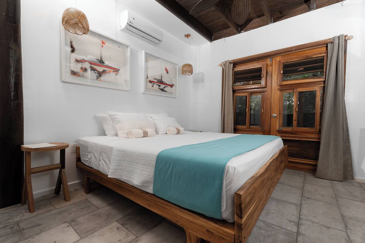 A bedroom with a wooden bed, white bedding, a teal throw blanket, a small wooden stool, and a window with curtains.
