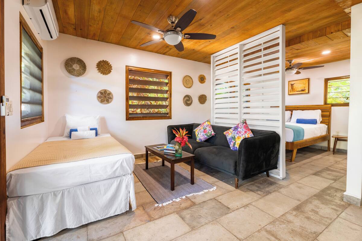 A room with a bed, a sofa, a coffee table with books and a vase, decorative wall plates, and a ceiling fan.