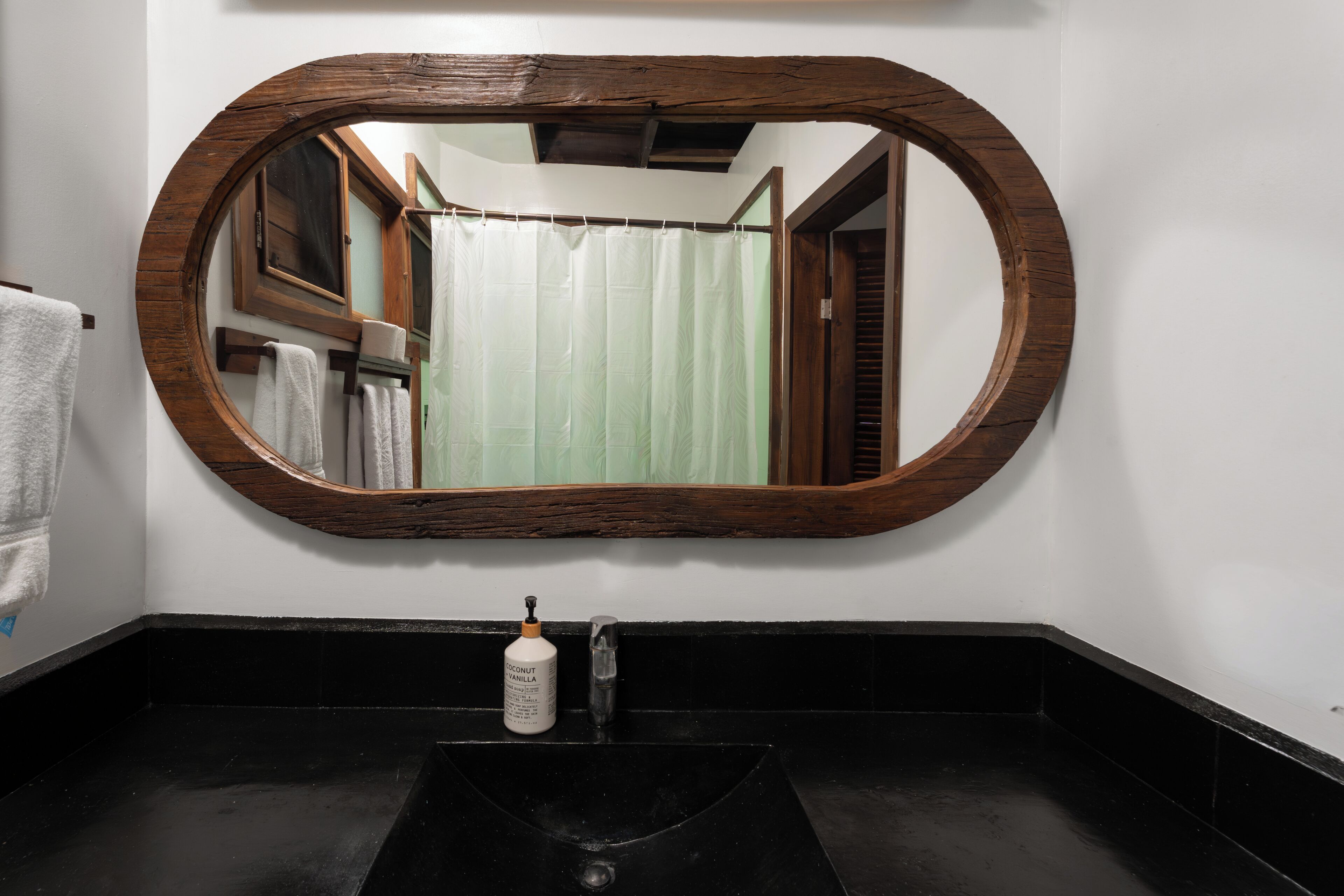 A bathroom with a large oval mirror, a black sink, and a bottle of coconut vanilla soap.