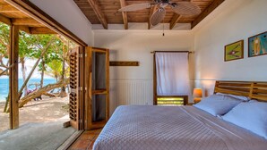 A bedroom with a bed, a ceiling fan, a window with a view of the beach, and a door leading to an outdoor area. - CAMP BAY LODGE (Roatan)