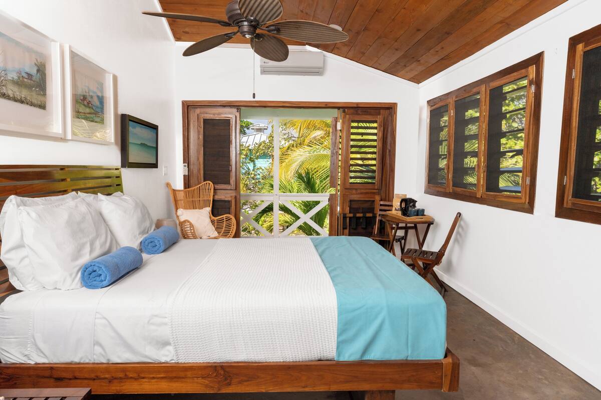 A bedroom with a wooden bed, a ceiling fan, and a view of a lush garden through a glass door.
