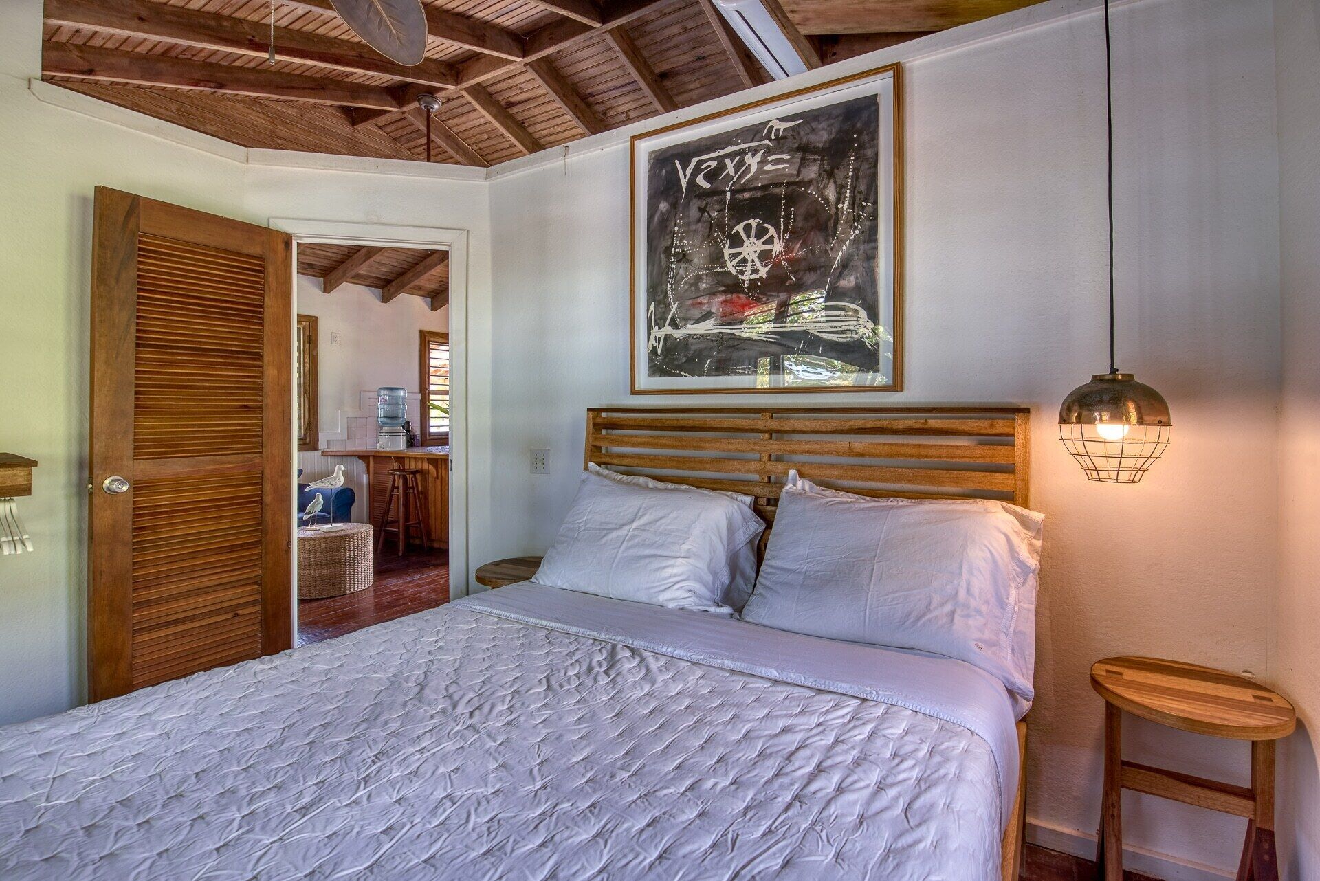 Un dormitorio con cabecera de madera, una cama grande con ropa de cama blanca, una lámpara colgante y un cuadro abstracto enmarcado en la pared.