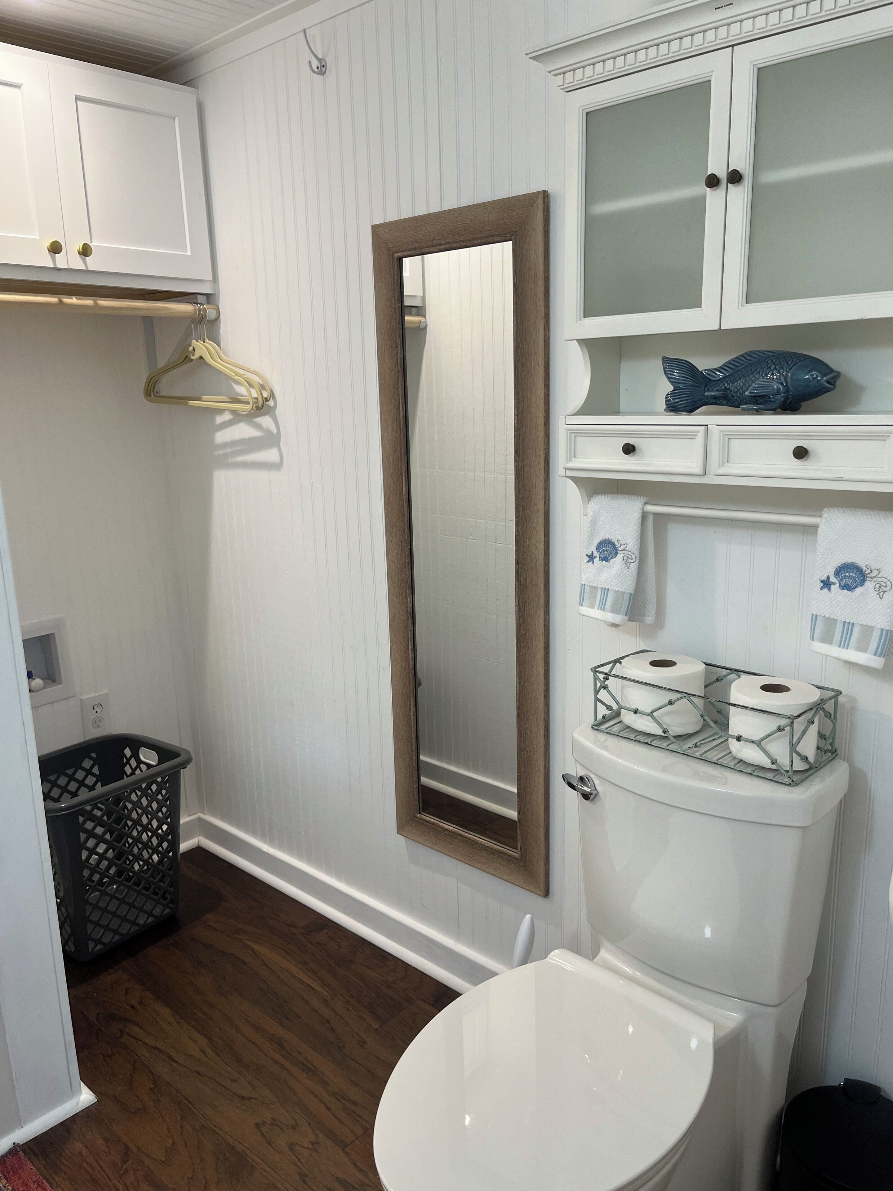 A bathroom with a toilet, a mirror, a hanging clothes rack, and a shelf with a decorative fish.