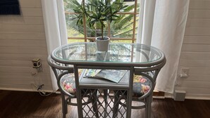 Une table avec un plateau en verre, agrémentée d’un coussin à motifs floraux, d’une plante en pot et d’un magazine, le tout placé devant un mur blanc avec une fenêtre.