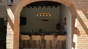 Zona de bar con un arco de ladrillo, cuatro taburetes de mimbre, una lámpara colgante y una barra con varios objetos.