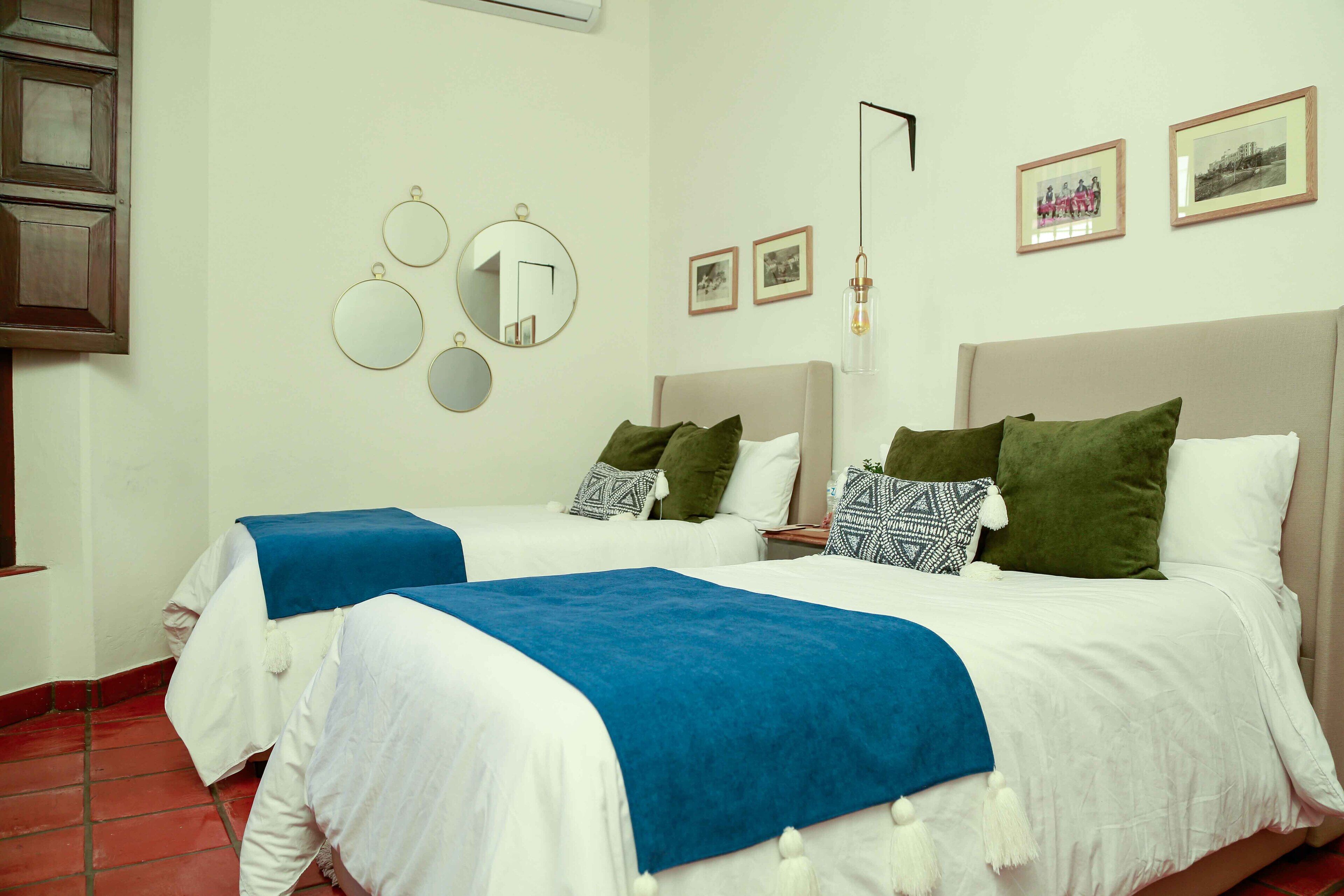 A hotel room with two beds, a red tiled floor, and a wall with three framed pictures and three round mirrors.