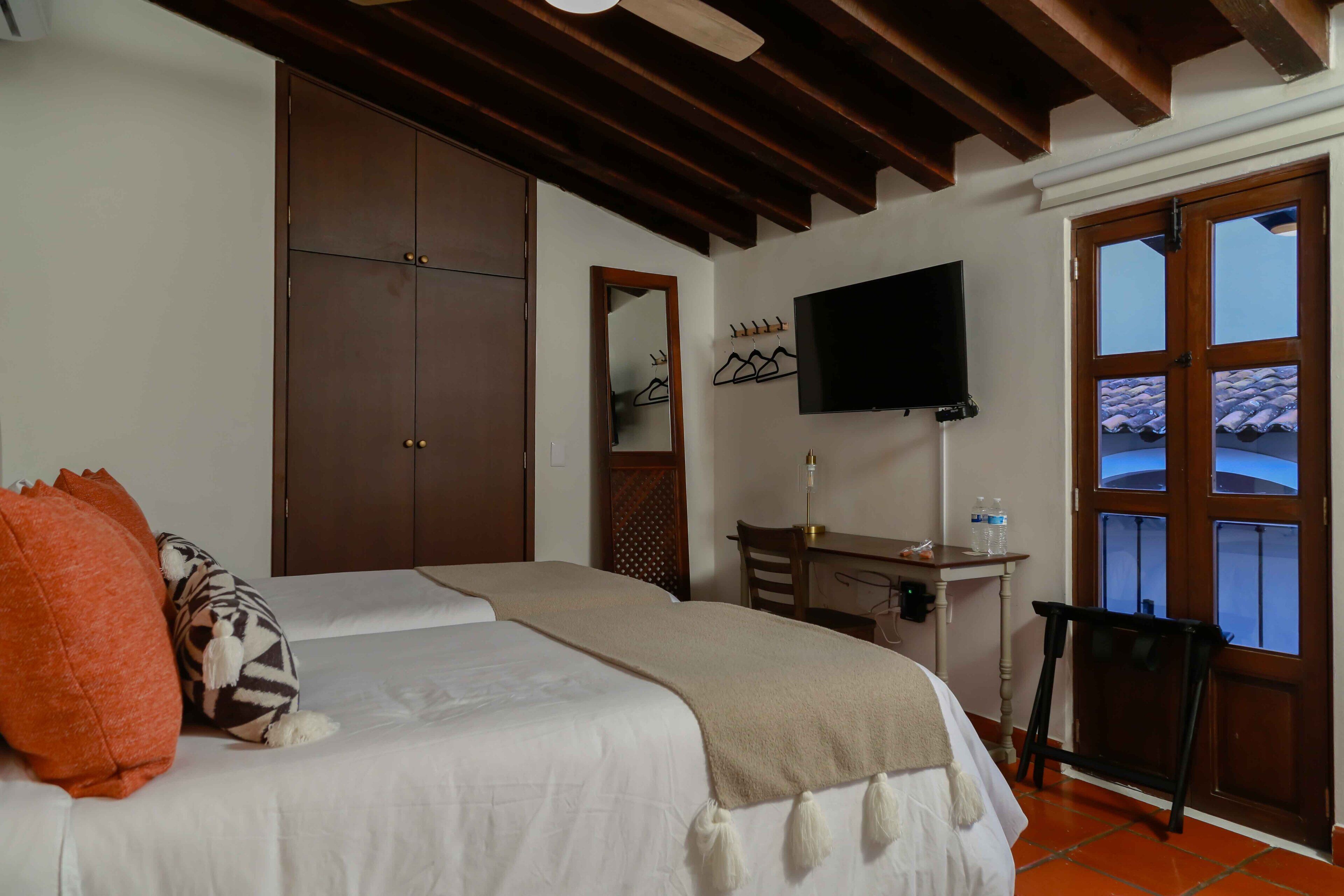 A bedroom with a bed, a wardrobe, a desk with a chair, a television, and a window with a view of the roof.