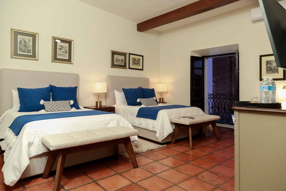 A hotel room with two beds, each with a white and blue bedspread, a bench, and a small table. There are framed pictures on the wall and a water dispenser on a cabinet.