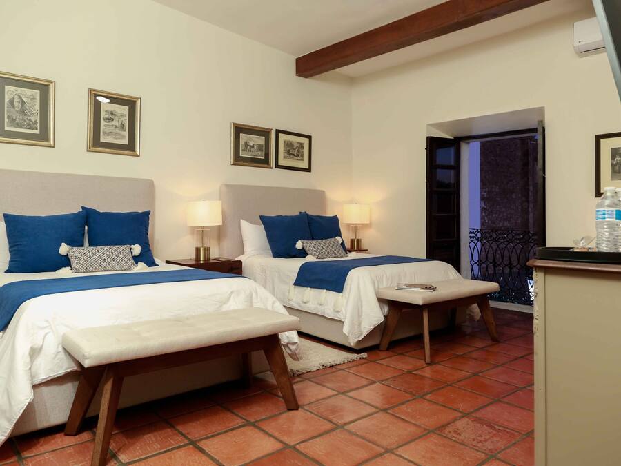 A hotel room with two beds, each with a white and blue bedspread, a bench, and a small table. There are framed pictures on the wall and a water dispenser on a cabinet.