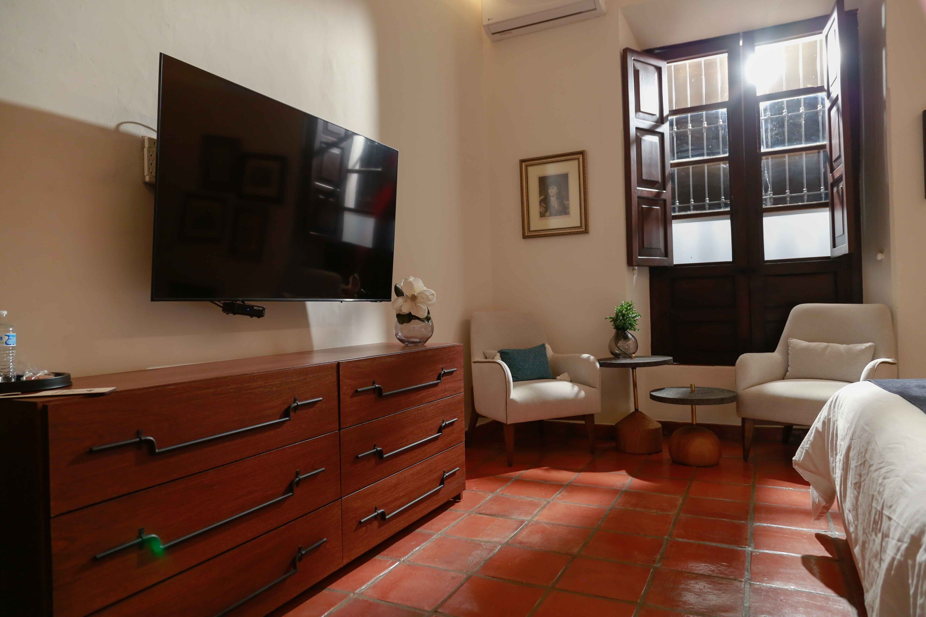 A hotel room with a TV mounted on the wall, a wooden dresser, a white armchair, a small round table with a plant, and a window with a view.