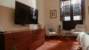 A hotel room with a TV mounted on the wall, a wooden dresser, a white armchair, a small round table with a plant, and a window with a view.