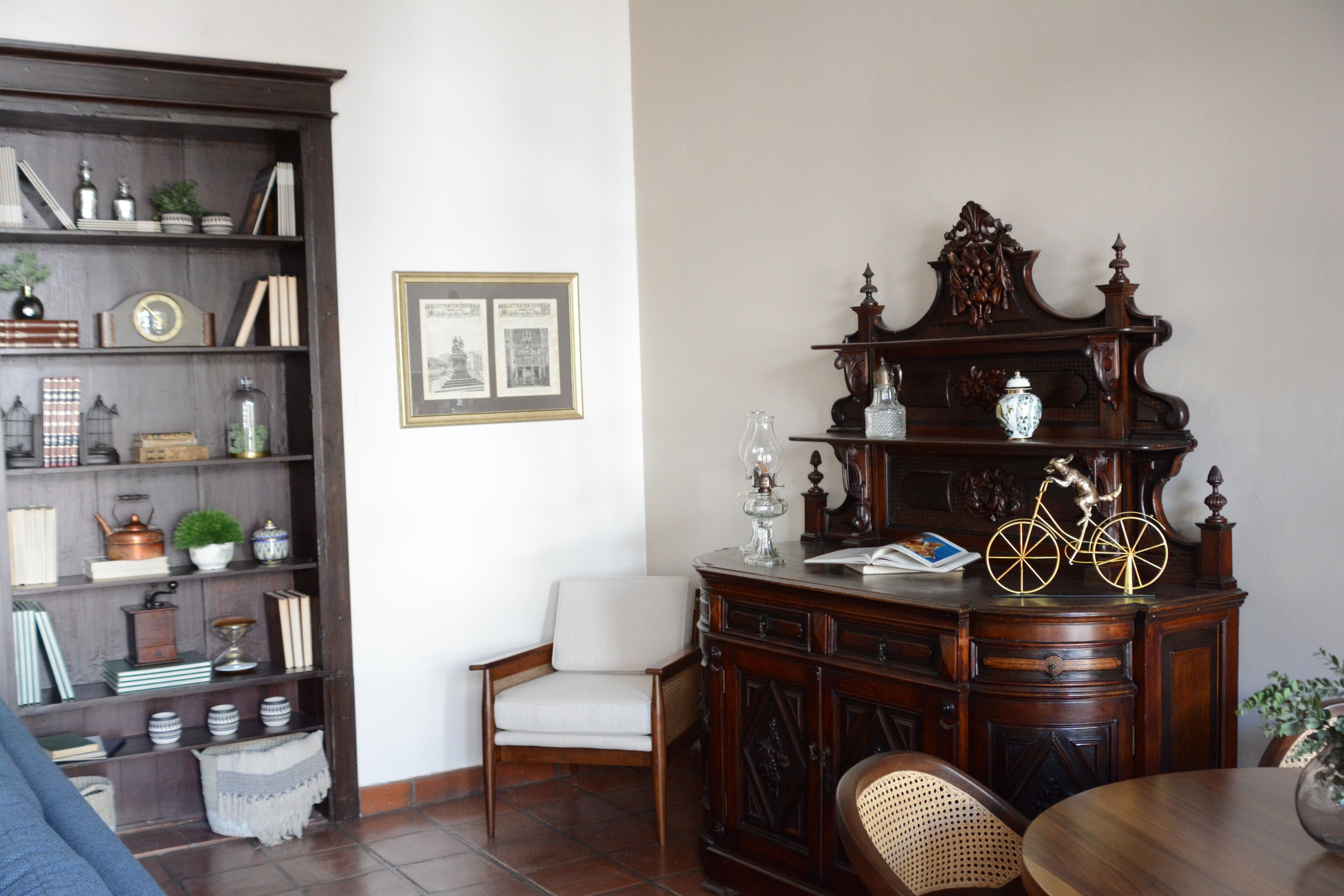 Un soggiorno con un mobile in legno scuro, una poltrona bianca e una sedia in vimini. Si nota una libreria piena di libri e oggetti decorativi.