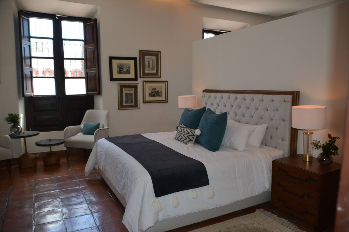 A bedroom with a large bed, two bedside tables, a chair, a window with shutters, and framed pictures on the wall.