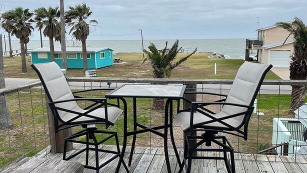 A wooden deck with two chairs and a table, overlooking a beach with palm trees and a small building.