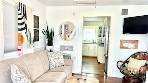 A living room with a sofa, a coffee table, a wicker chair, a potted plant, a mirror, and a kitchen in the background.