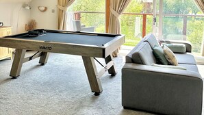 Una mesa de billar en una habitación con techo inclinado, un sofá y ventanas amplias con cortinas.