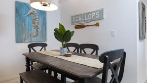 A dining area with a wooden table, four chairs, a potted plant, a decorative wall sign, and a seahorse painting. - Two 2BD/2BA 1st floor Condos for Group/Family/Sports Team Vacation (Gulf Shores)