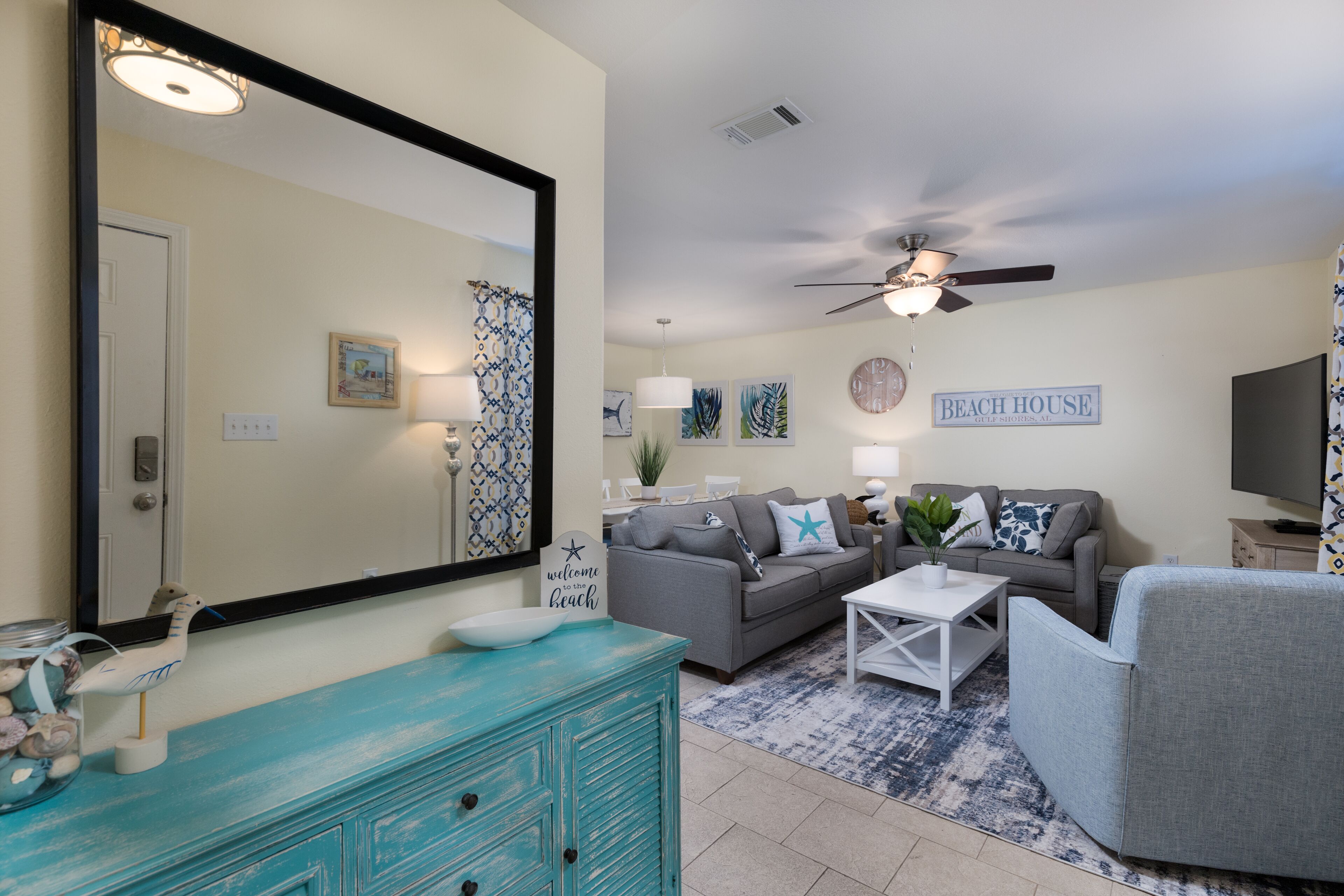 Un salon avec un canapé gris, une commode turquoise, un ventilateur de plafond et un panneau indiquant « MAISON DE PLAGE ».