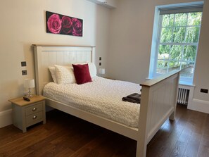 A bedroom with a white bed, a red pillow, a nightstand with a lamp, and a window with a view of greenery.