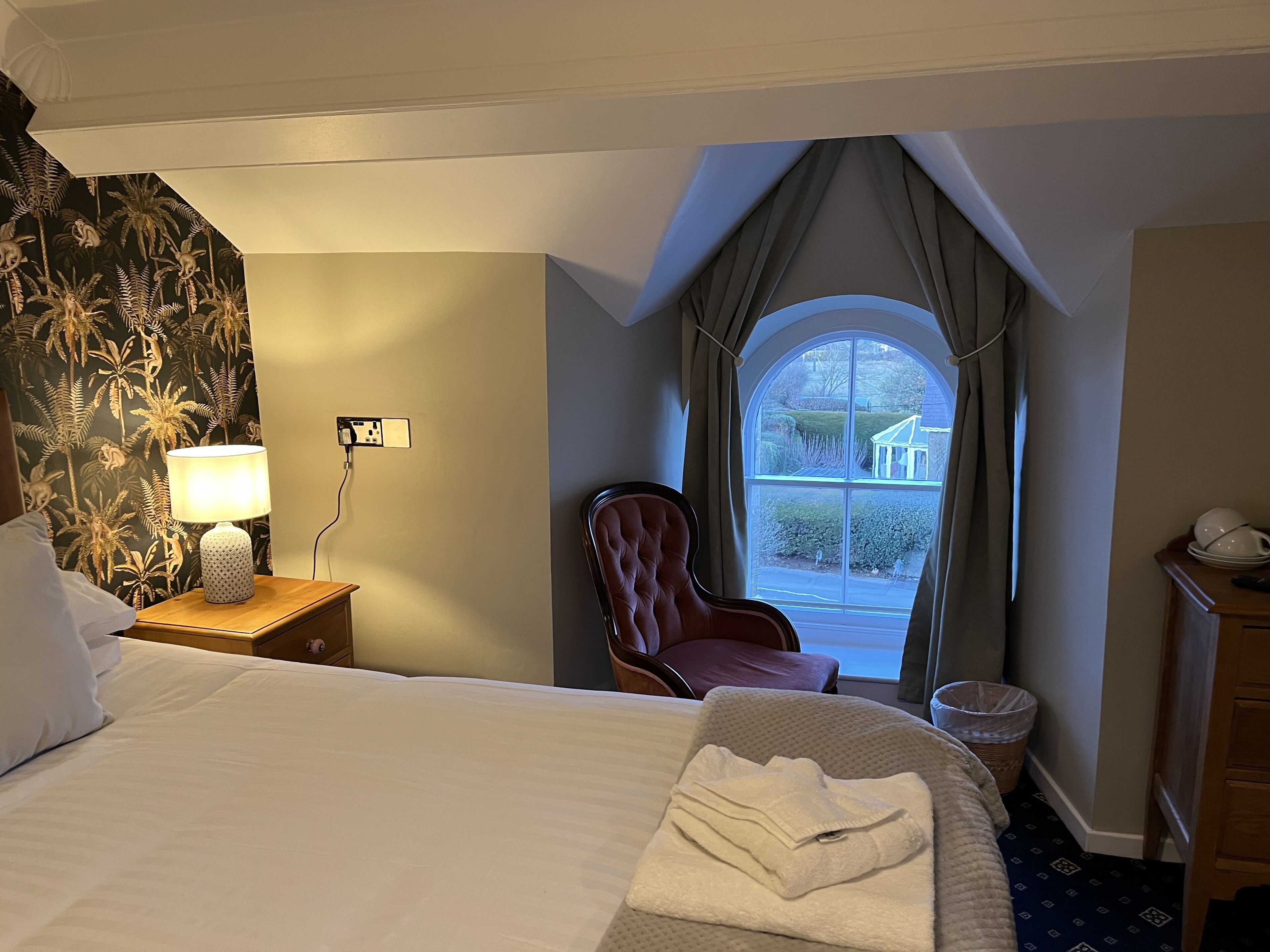 Ein Hotelzimmer mit Bett, Sessel, Lampe, einem Fenster mit Vorhängen und Blick ins Freie.