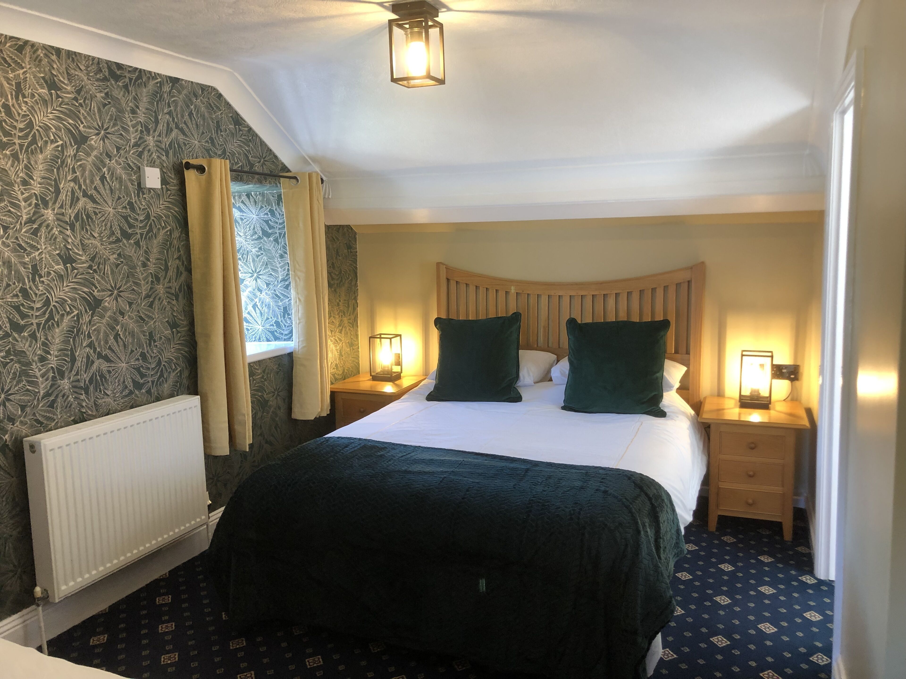 A hotel room with a bed, two bedside tables with lamps, a window with curtains, a radiator, and patterned wallpaper.