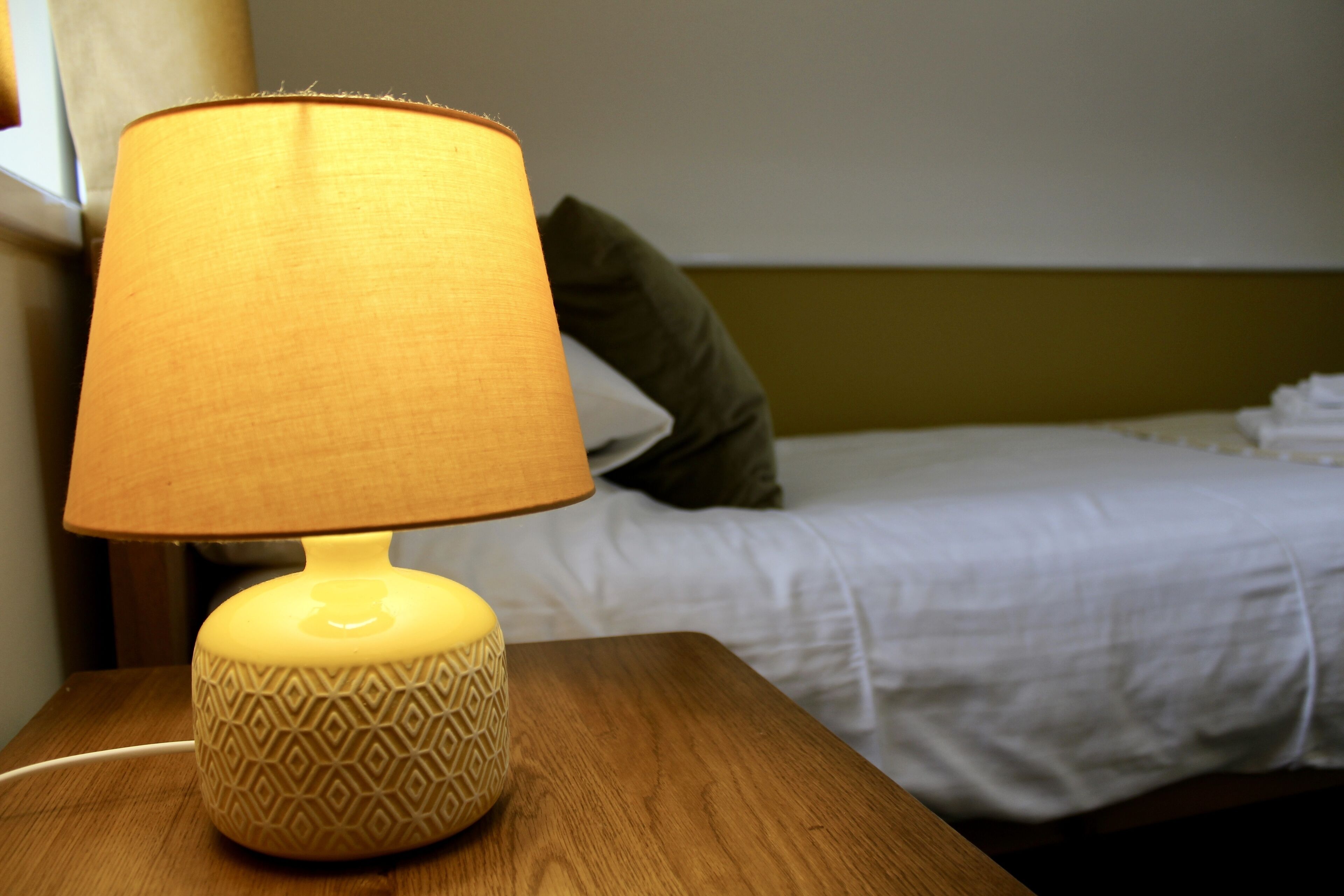 A bedside table with a patterned lamp, a bed with white bedding, and a green pillow.