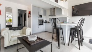 Ein modernes Wohnzimmer mit einer Couch, einem Couchtisch und einem Fernseher. Es gibt eine Küchenzeile mit einer Mikrowelle, einem Backofen und einer Bar mit Hockern.