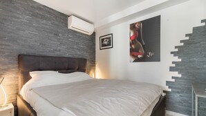 Ein modernes Schlafzimmer mit dunklem Kopfteil, einem großen Bett mit weißen Leinen und einer Wand-Klimaanlage. Darüber am Bett hängt ein gerahmtes Bild.