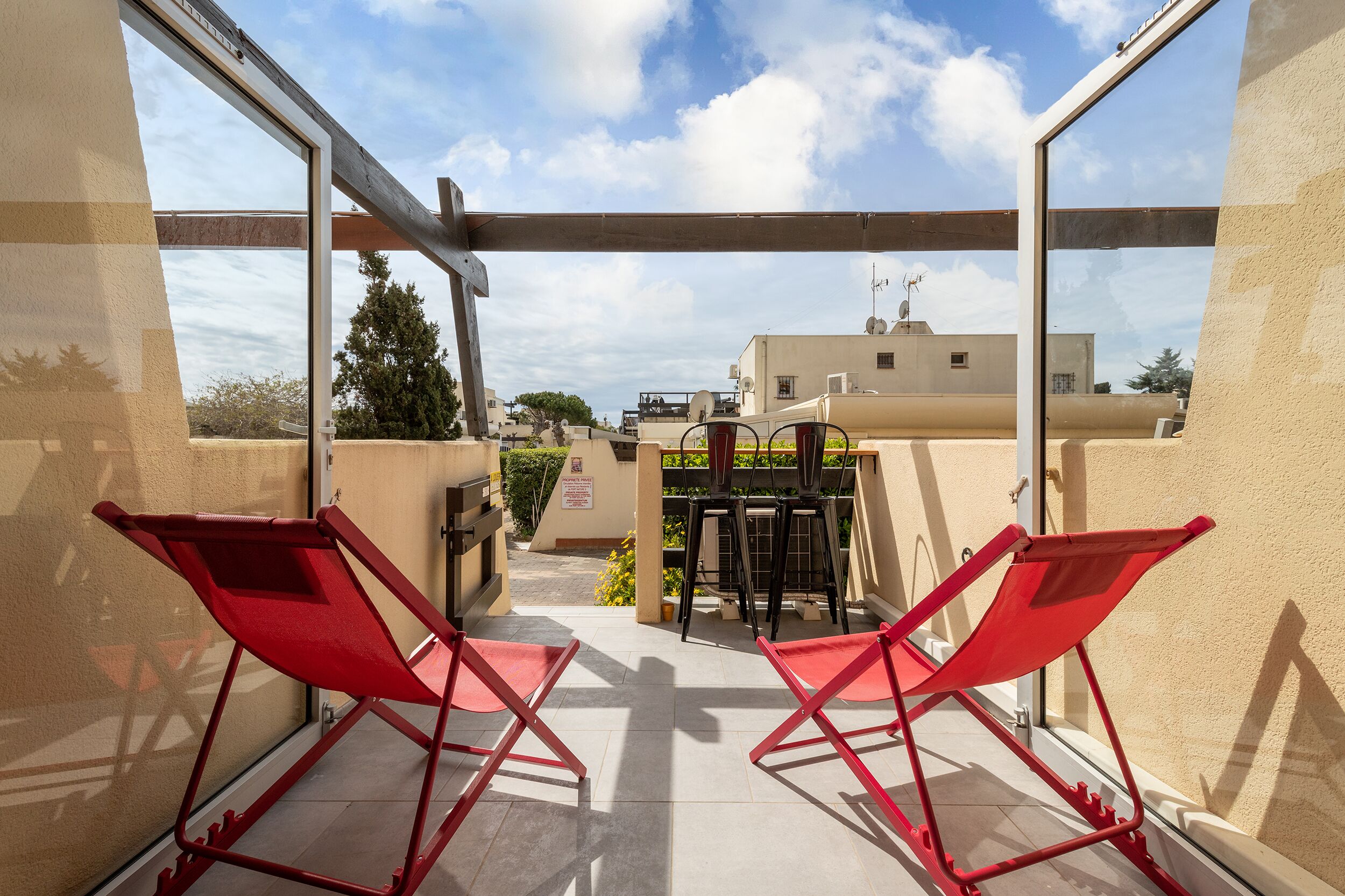 Un balcone con due sedie rosse pieghevoli, una porta vetrata e vista su edifici e alberi sullo sfondo.