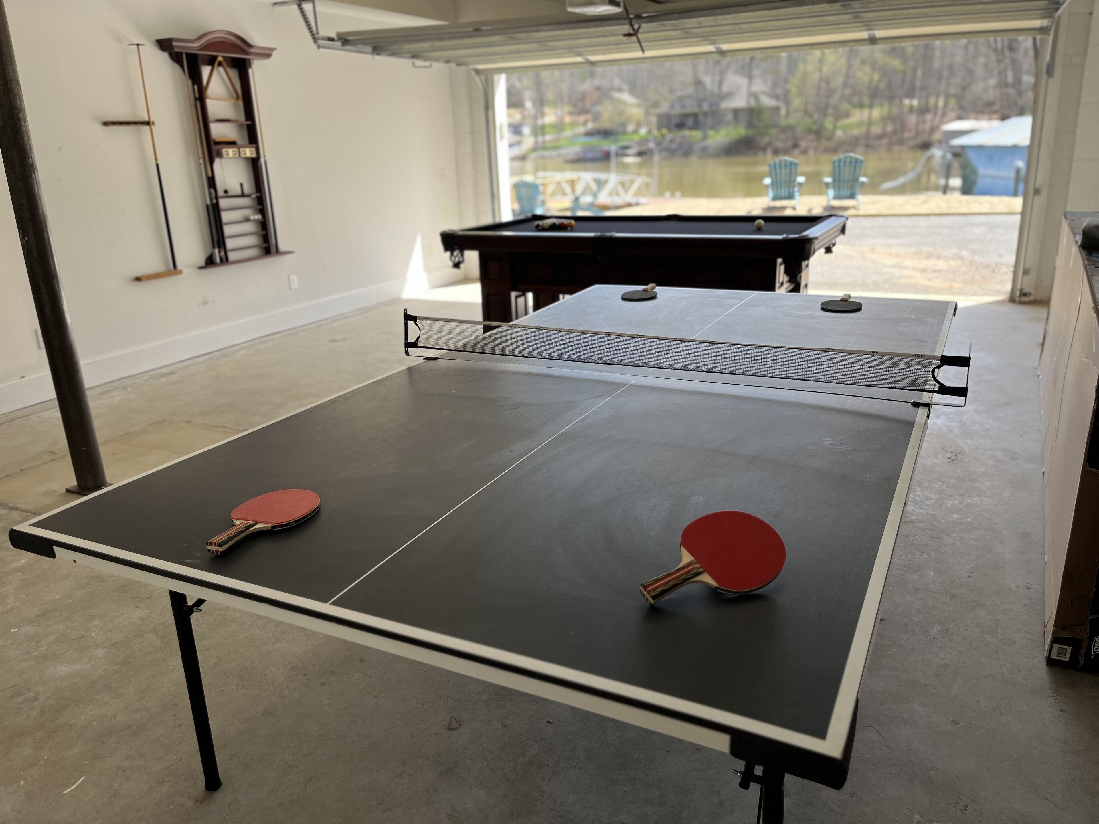 Une table de ping-pong avec deux raquettes, installée dans un espace intérieur offrant une vue sur un lac et un quai.