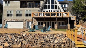 A two-story house with a stone facade, a large garage, and a covered outdoor seating area with Adirondack chairs. - The Chalet on Lake Wylie, 5000 sq ft, Sleeps16, Pet Friendly, Close to Charlotte (Belmont)