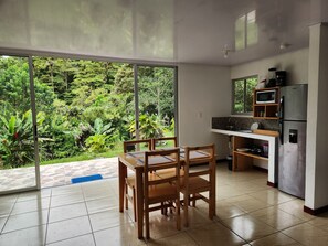 A modern kitchen with a built-in microwave, refrigerator, and sink. There is a dining area with a table and chairs, and large windows offering a view of lush greenery. - Selvática del Toro  (Toro Amarillo)