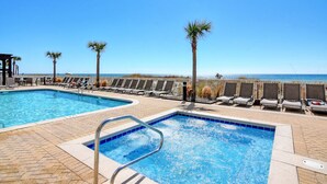A pool area with two pools, lounge chairs, palm trees, and a view of the ocean. - Ocean Villa #2106 by Resort Collection (Panama City Beach)