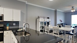 A modern kitchen with white cabinets, a black countertop, and a dining area with white chairs and a table. - Ocean Villa #2106 by Resort Collection (Panama City Beach)