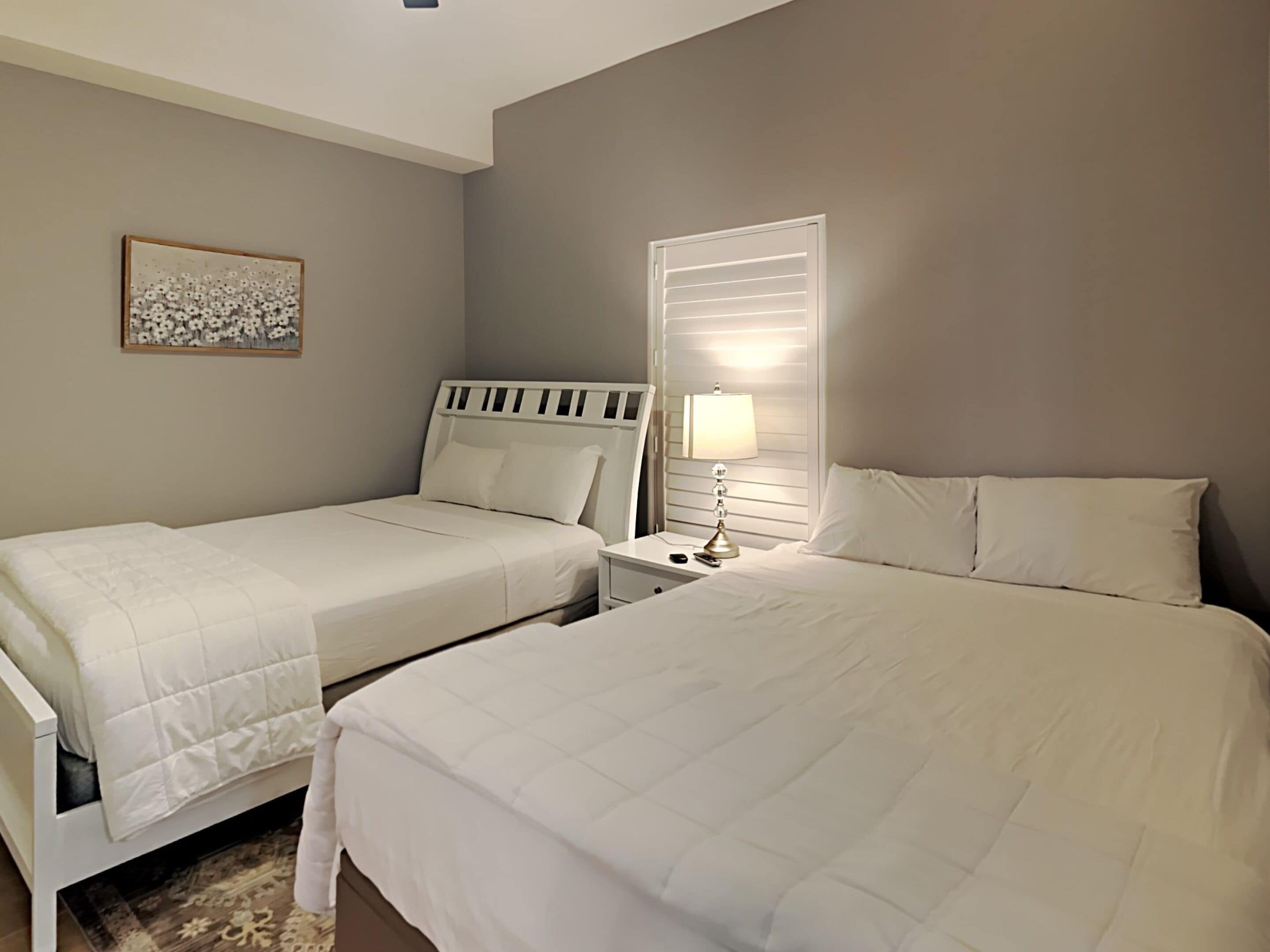 Une chambre avec deux lits, une table de chevet avec une lampe, une fenêtre avec des stores et une œuvre d’art florale accrochée au mur.