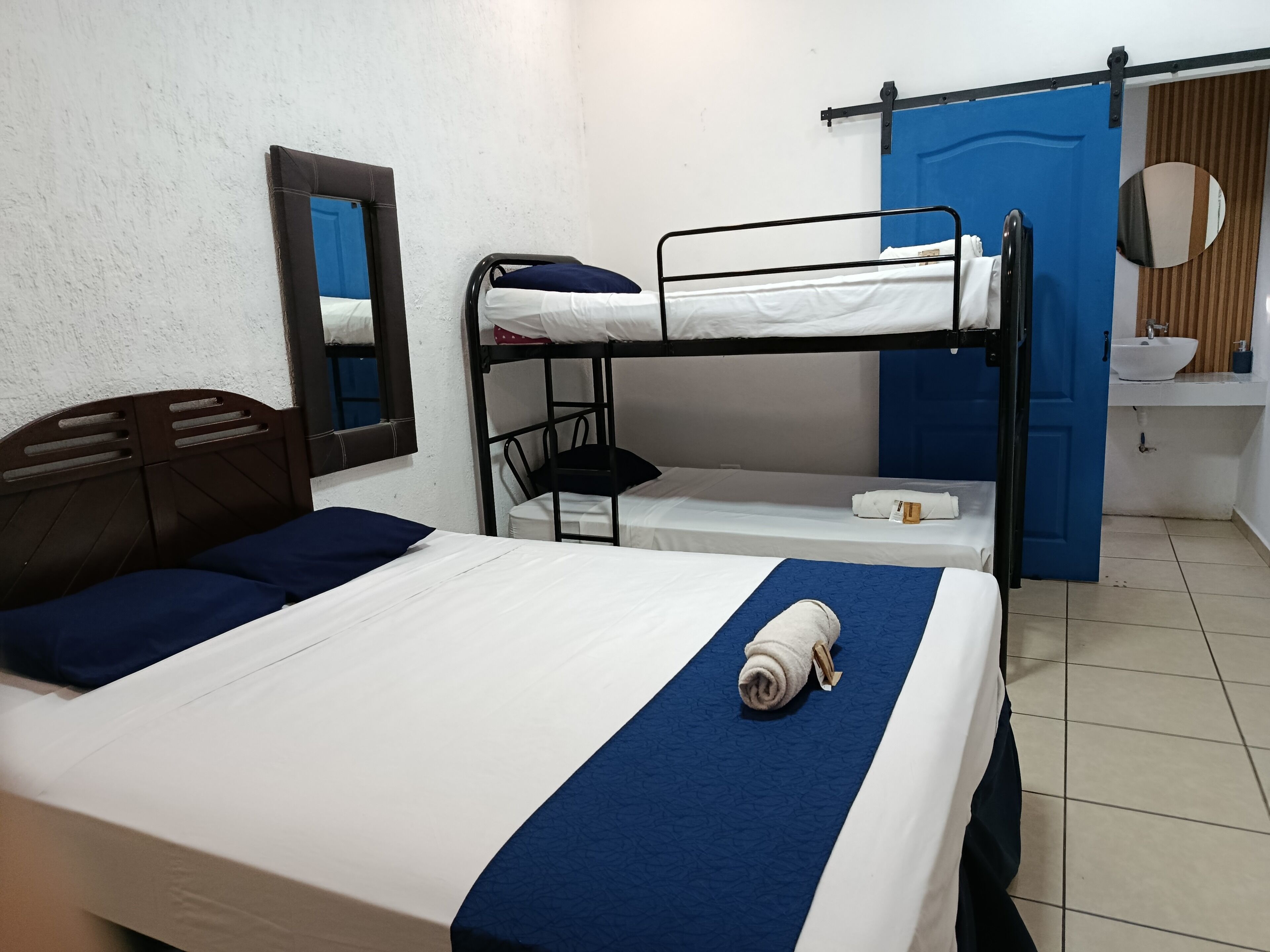 Une chambre avec un lit superposé, un lit simple, un miroir, un lavabo et une porte bleue.