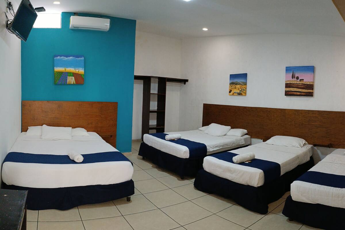 Een hotelkamer met vier eenpersoonsbedden, een houten hoofdbord, een blauwe accentmuur en een schilderij van een woestijnlandschap.