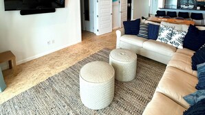 Un salon moderne doté d’un grand téléviseur à écran plat, d’un canapé, de poufs et d’un coin cuisine à l’arrière-plan.