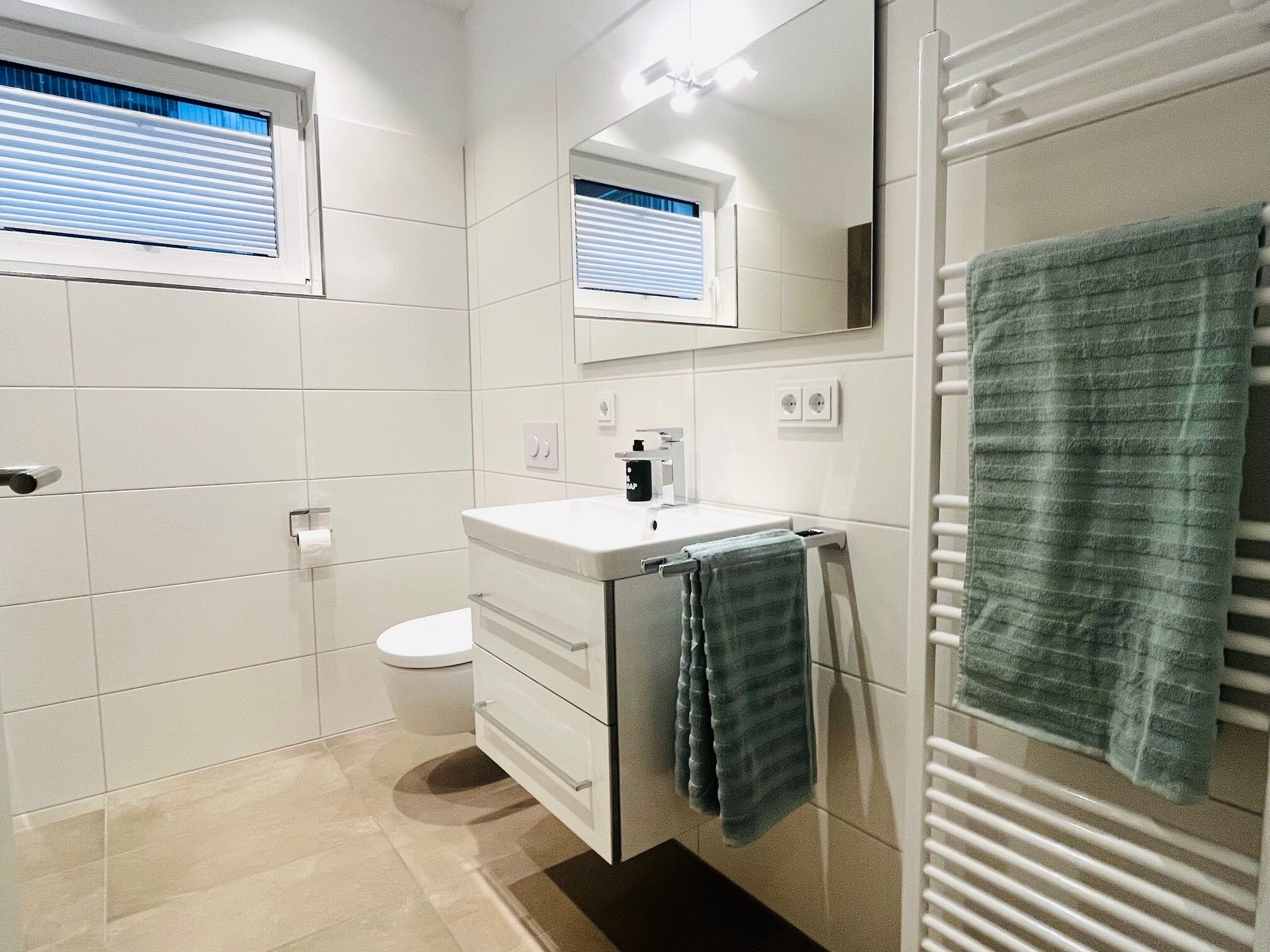 A modern bathroom with white tiles, a toilet, a sink with a mirror above it, and a towel rack with a towel.
