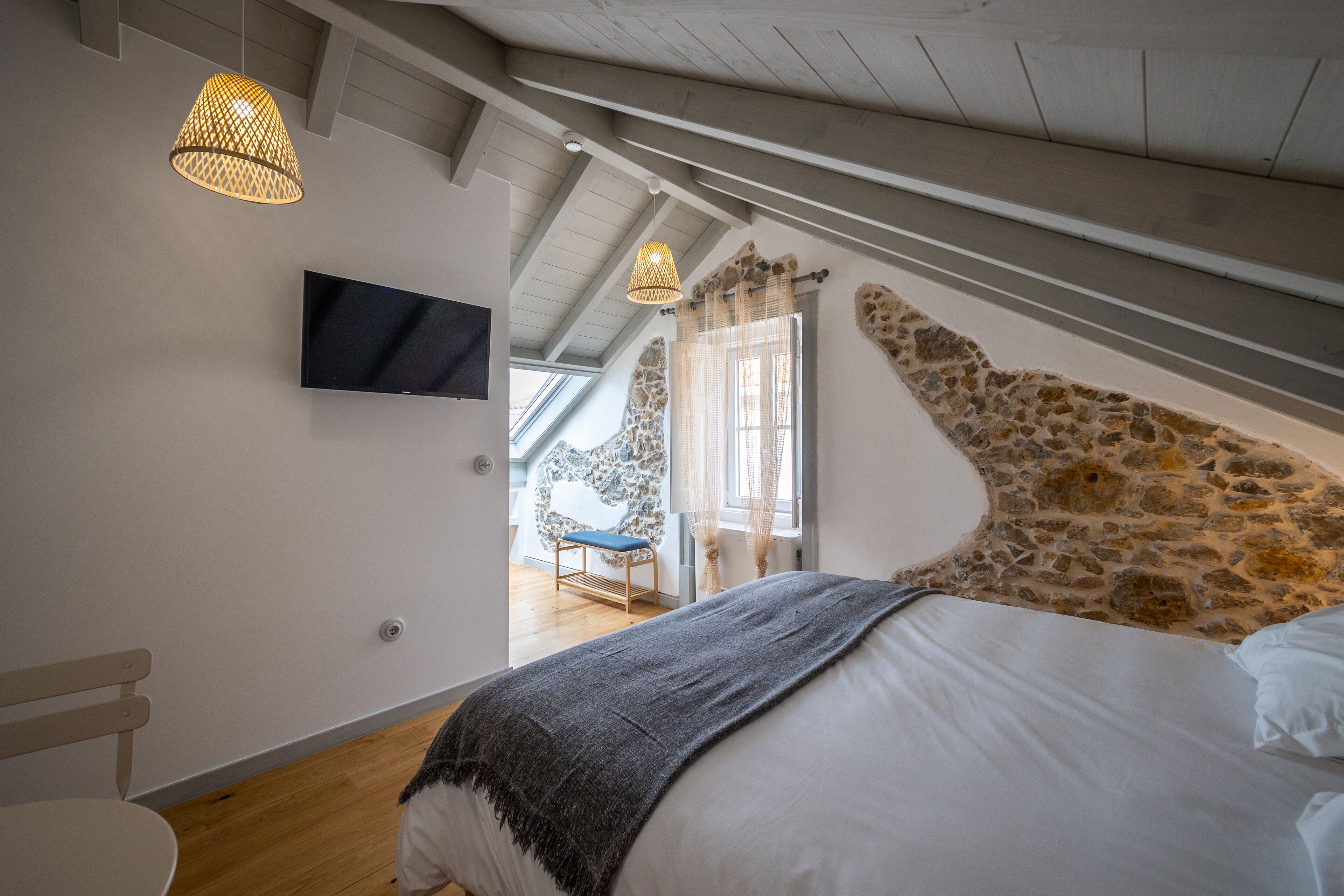 Une chambre avec un mur d’accent en pierre, un lit avec du linge de lit blanc, un plaid gris et un téléviseur fixé au mur.