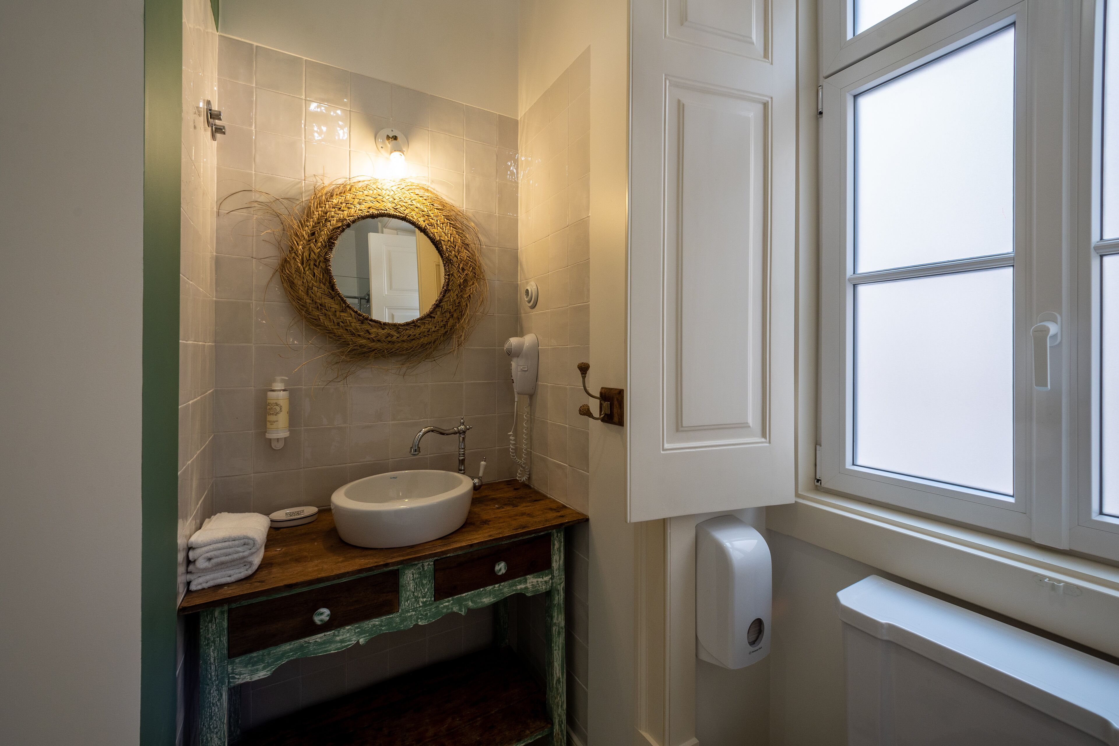 A bathroom with a round mirror, white sink, wooden vanity, and a window with a view.