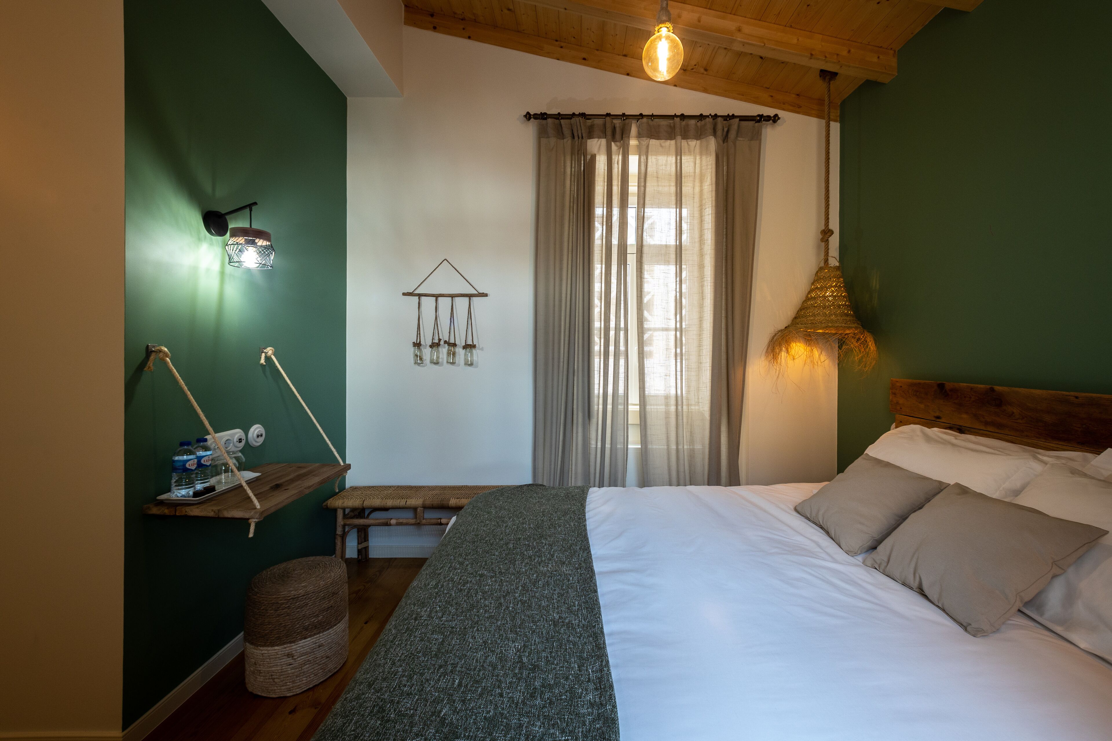 A bedroom with a bed, a hanging light fixture, a shelf with items, and a window with curtains.