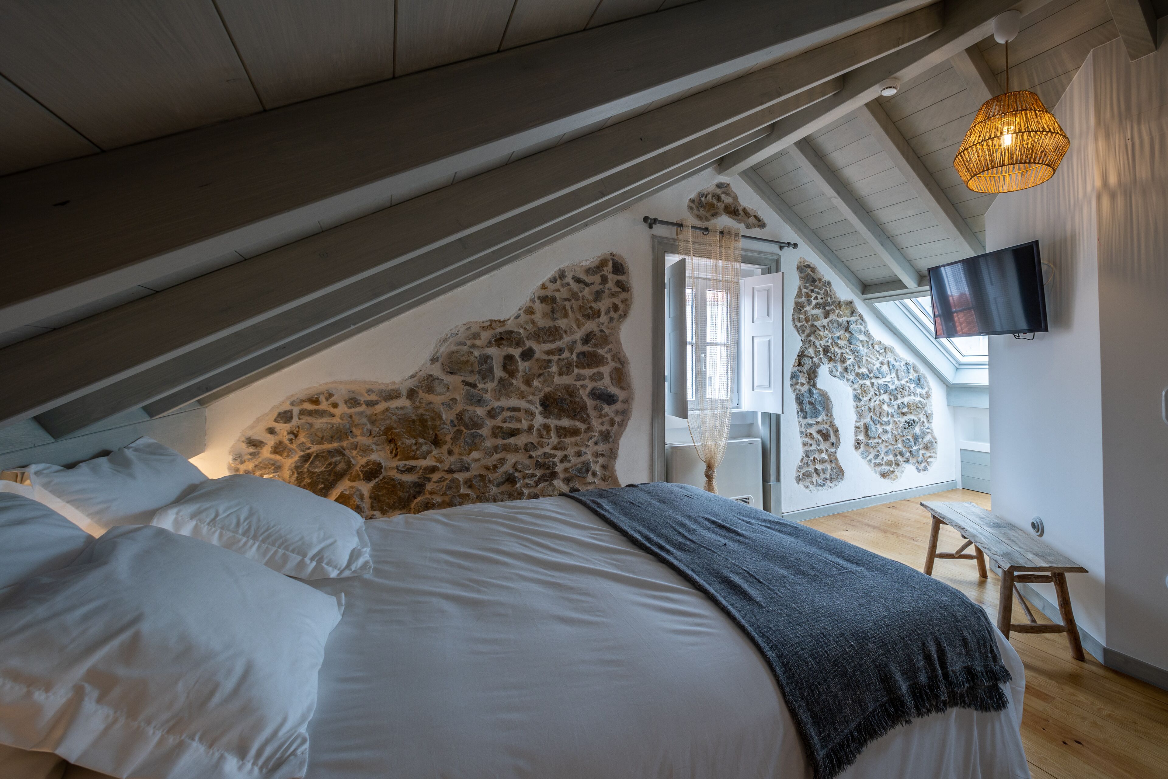 Une chambre avec un mur en pierre, un lit recouvert de draps blancs, un banc en bois et un téléviseur fixé au mur.
