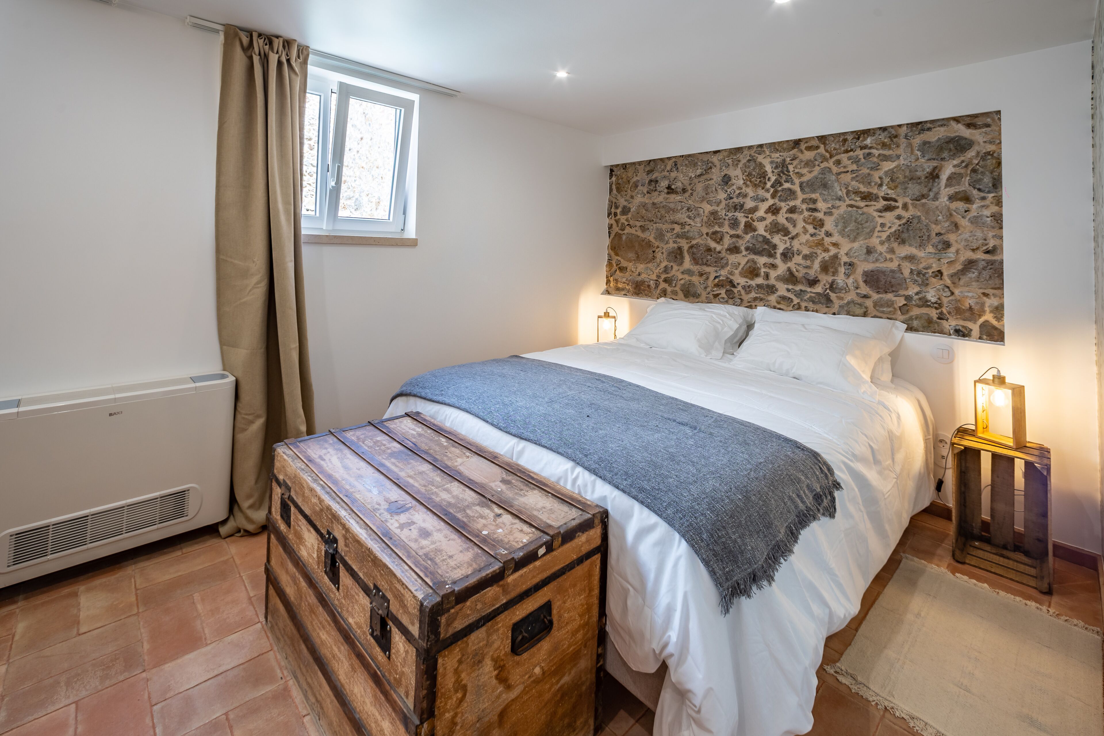 A bedroom with a stone wall, a bed with white linens, a wooden chest, a heater, and a nightstand with a lamp.