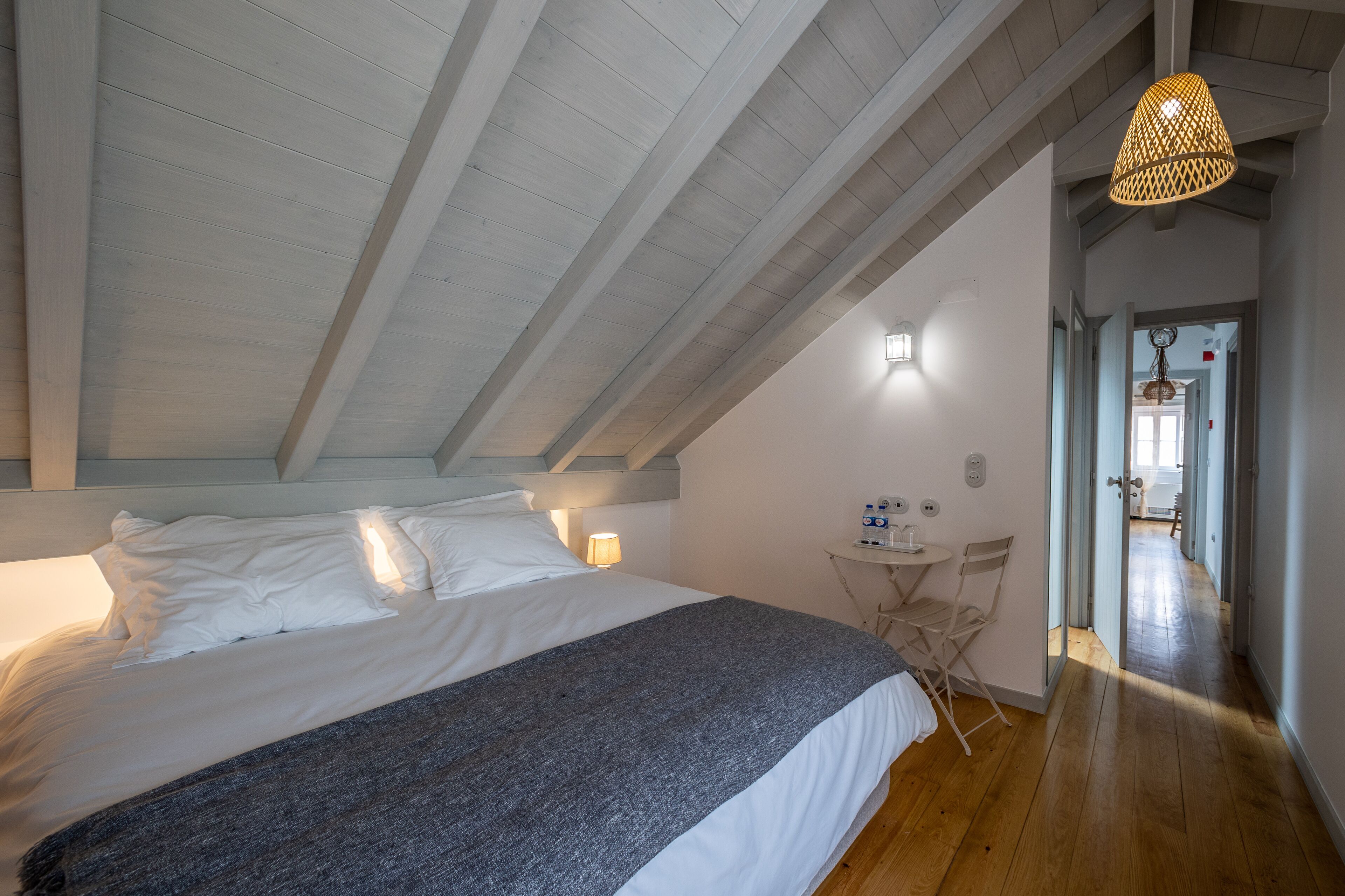 Un dormitorio con una cama grande, una mesita pequeña y una silla. El techo tiene vigas de madera y una lámpara colgante.