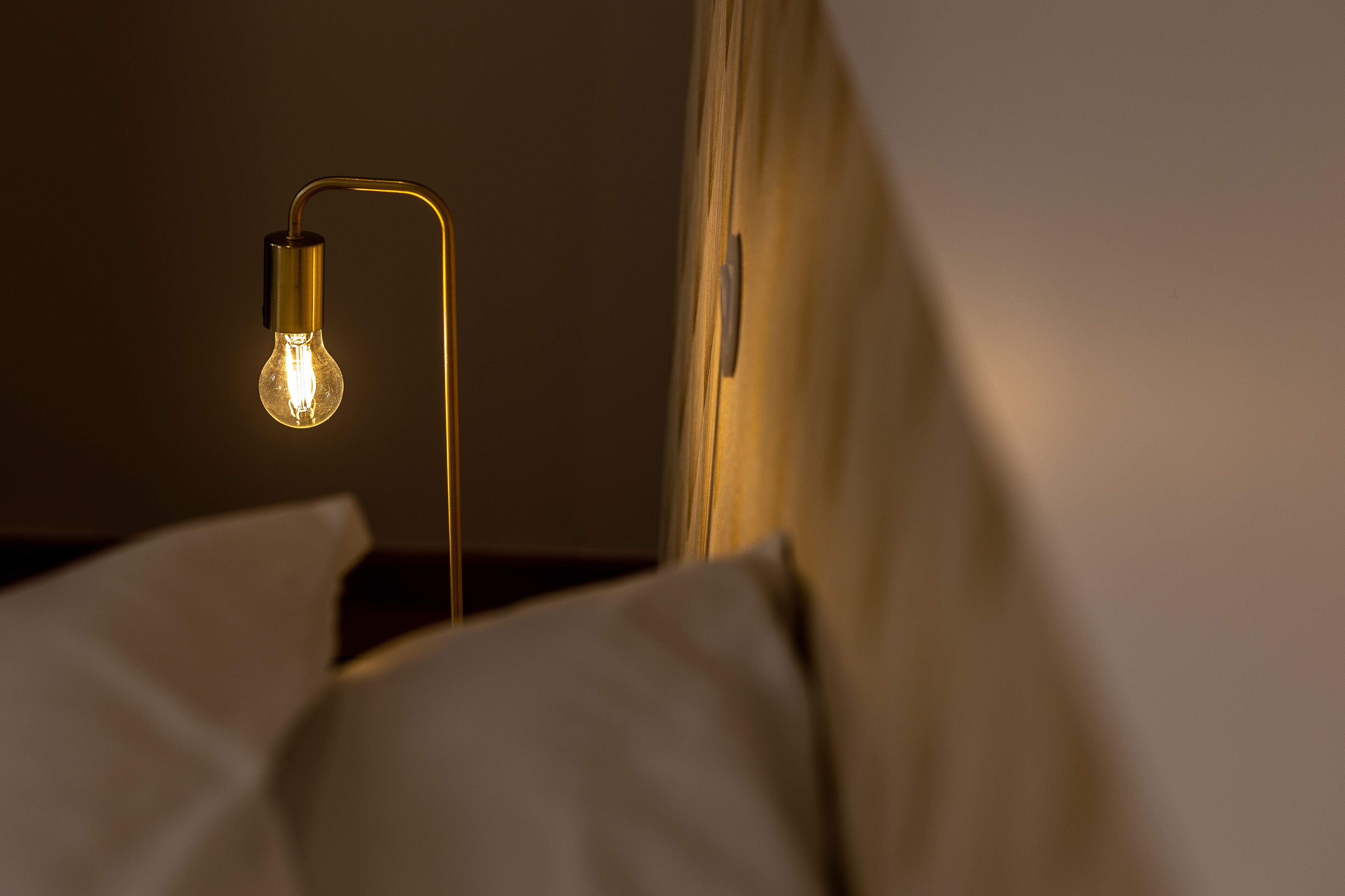 Een enkele gloeilamp hangt aan een gebogen metalen fitting, met een onscherpe achtergrond die een kamer doet vermoeden.