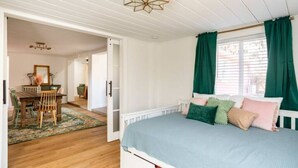 A bedroom with a bed, green curtains, a white ceiling, and a dining area with a table and chairs. - Kin Cottage Accord (Accord)
