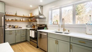 A modern kitchen with light green cabinets, stainless steel appliances, and a large window providing a view of the outdoors. - Kin Cottage Accord (Accord)
