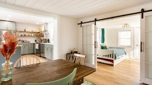 A modern kitchen with white cabinets, a dining table with chairs, and a sliding door leading to a bedroom with a bed and a window. - Kin Cottage Accord (Accord)